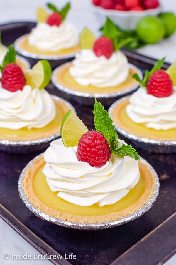 A dark sheet pan with 6 mini key lime pies topped with whipped cream and raspberries.