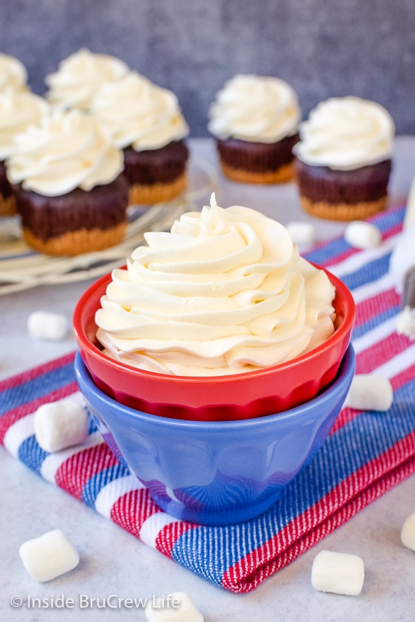 A stack of red and blue bowls filled with a swirl of marshmallow frosting and frosted cupcakes behind it.
