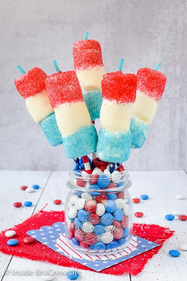 A clear jar filled with red white and blue m&m candies with five firecracker marshmallow pops in it.
