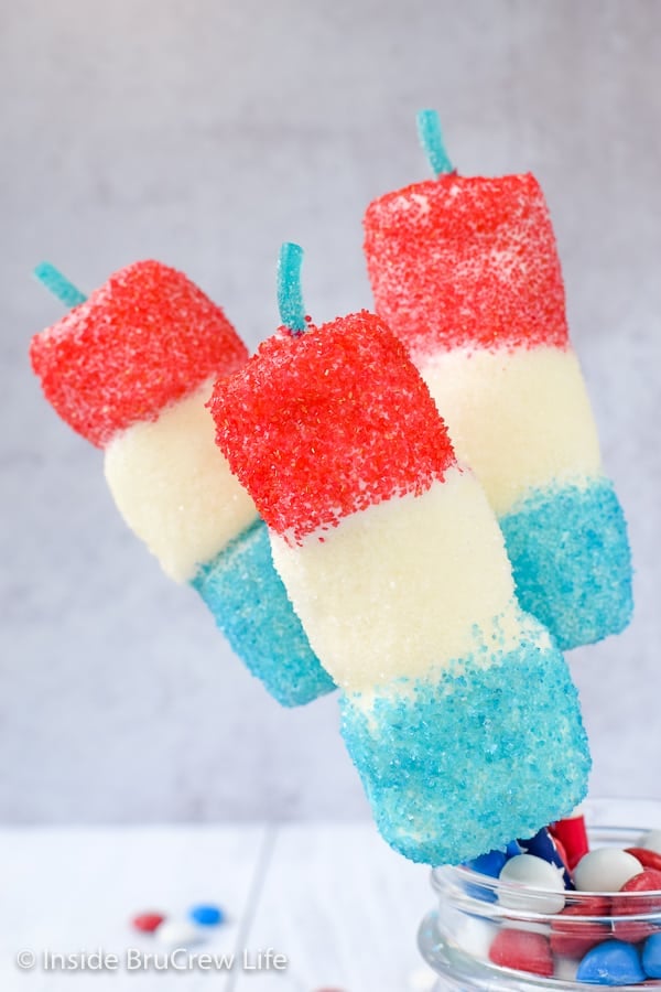 Three marshmallow firecracker pops sticking out of a jar of M&M's.