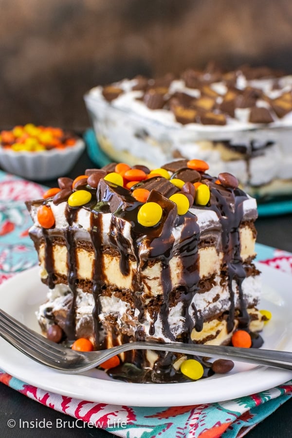 A white plate with a slice of Reese's Ice Cream Cake drizzled with chocolate syrup and topped with Reese's Pieces.