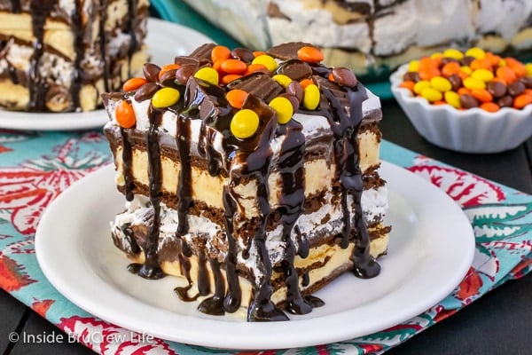 A white plate with a piece of Reese's ice cream cake topped with chocolate syrup and Reese's candies.