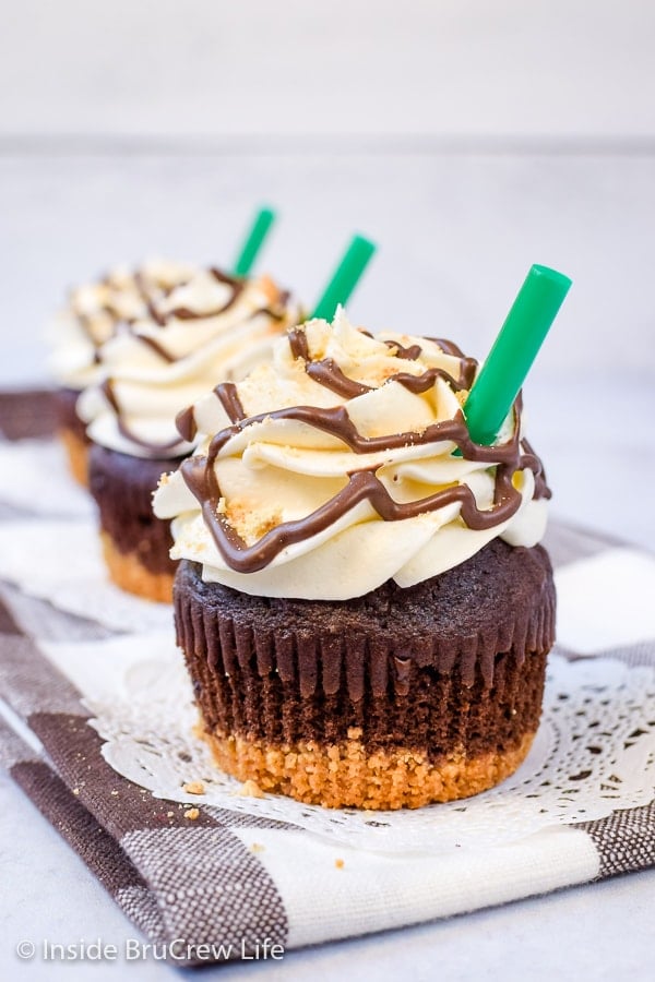 Three chocolate cupcakes topped with marshmallow frosting, chocolate drizzles, and a green straw on a white board.