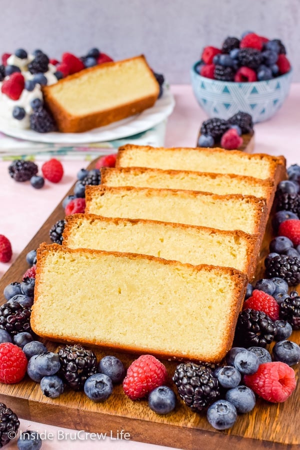 A loaf of vanilla pound cake sliced and arranged on a wooden tray with fresh berries all around it.