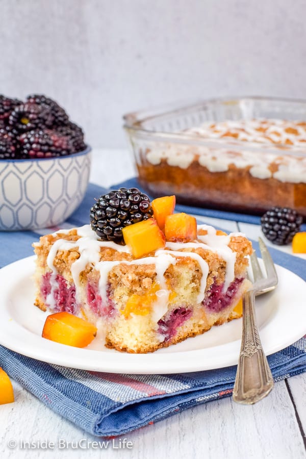 A white plate on a blue towel with a piece of peach coffee cake topped with blackberries on top.