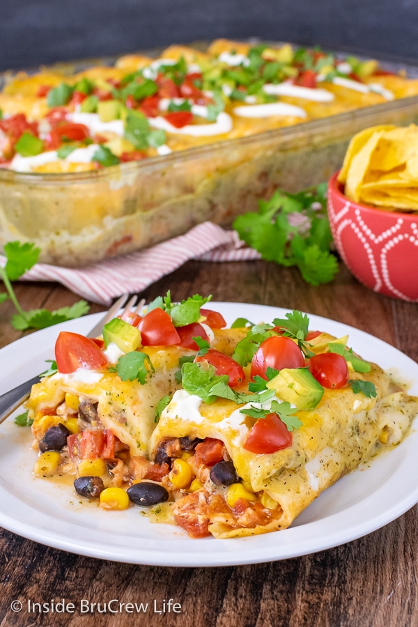 Two chicken enchiladas halves on a white plate topped with sour cream, tomatoes, and avocados.