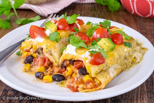 A chicken enchilada cut in half on a white plate and topped with sour cream, avocado, and tomatoes.