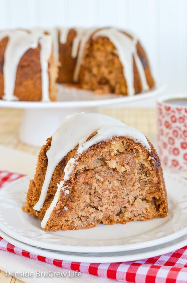 A white plate with a slice of apple bundt cake drizzled with apple cider glaze on top and the cake behind it.