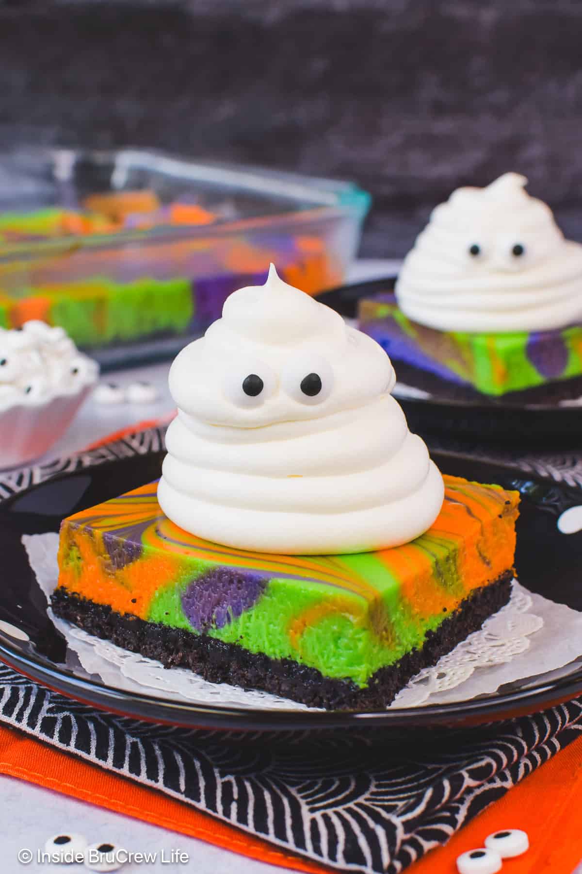 A square of halloween cheesecake topped with a whipped cream ghost on a black plate.