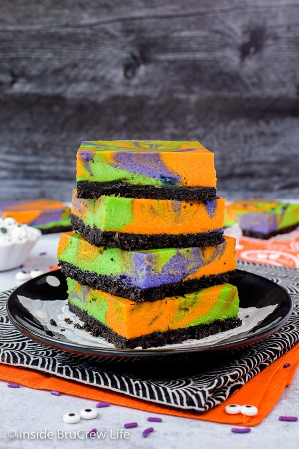 A black plate with a stack of four halloween swirled cheesecake bars with an Oreo crust on it.