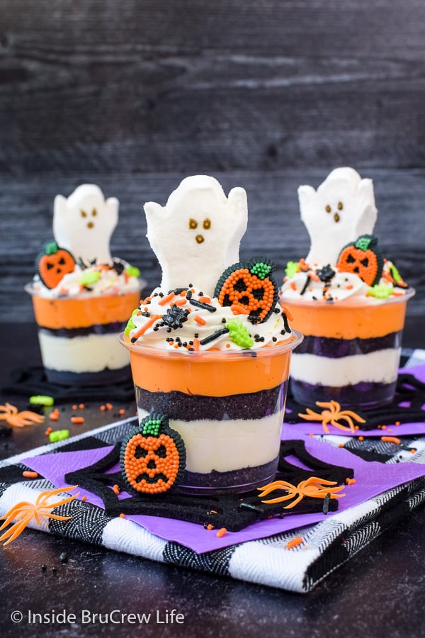 Three clear cups with orange and white cheesecake layers, Oreo cookie crumbs, and marshmallow ghosts on top.