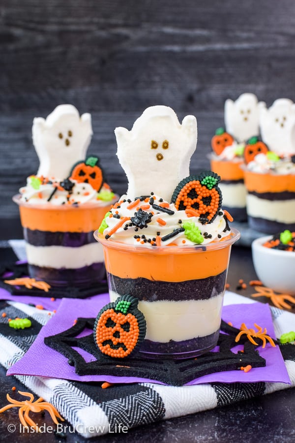Clear cups with layers of Oreo crumbs, orange and white cheesecake, and marshmallow ghosts on top.
