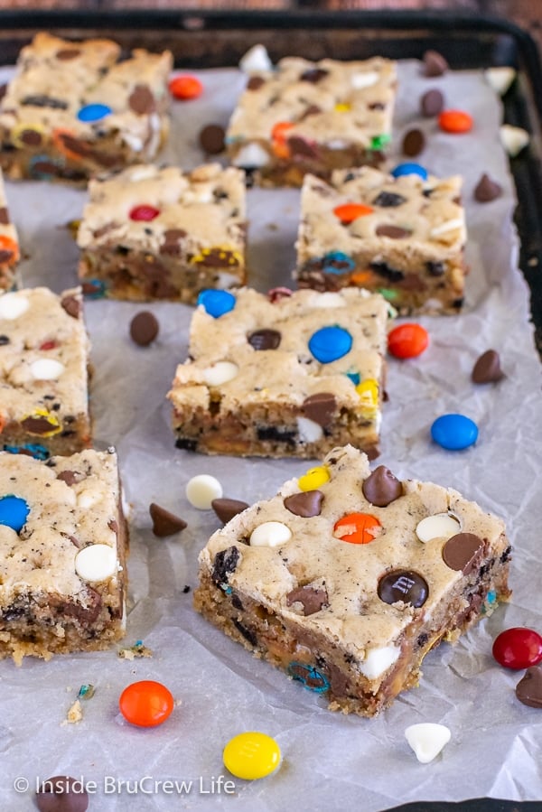 A sheet pan with parchment paper on it and squares of loaded blonde brownies and candy scattered on it