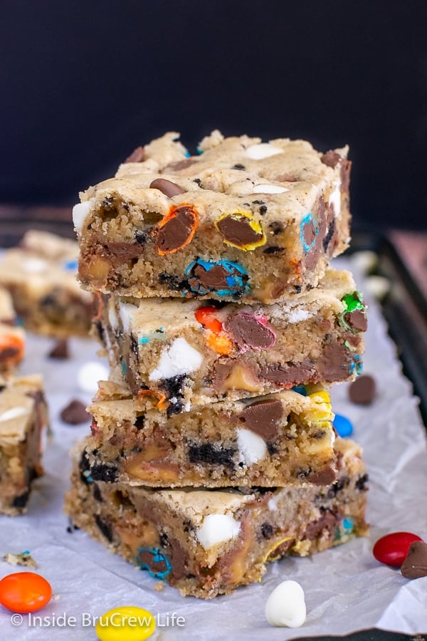 Four blonde brownies loaded with cookies, chocolate, and candy stacked on top of each other with candy around them