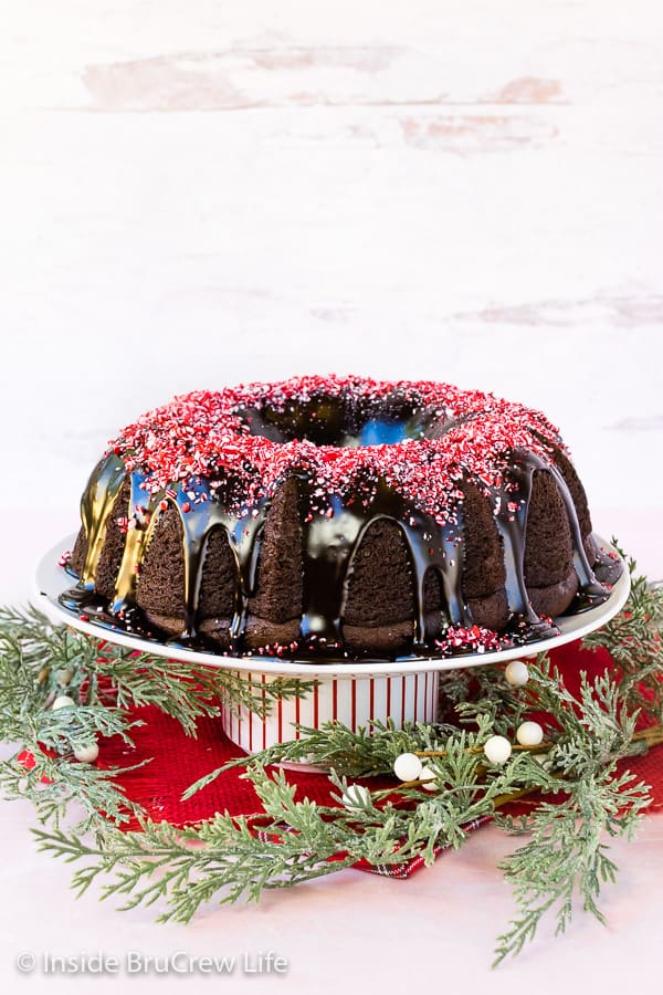 A white cake plate with a full chocolate bundt cake topped with ganache and peppermint bits.