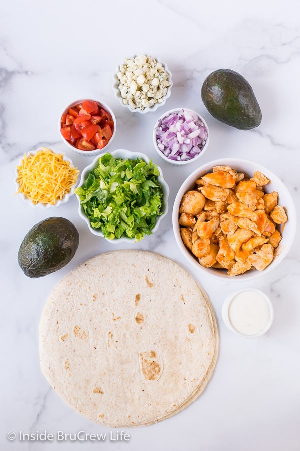 Tortillas and bowls of ingredients to make buffalo chicken wraps on a white board.