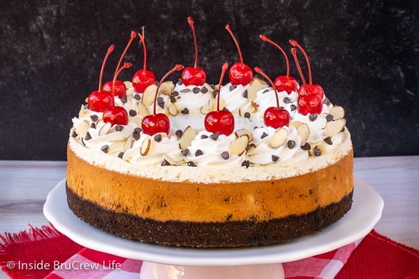 A full cheesecake topped with whipped cream and cherries on a white cake plate.