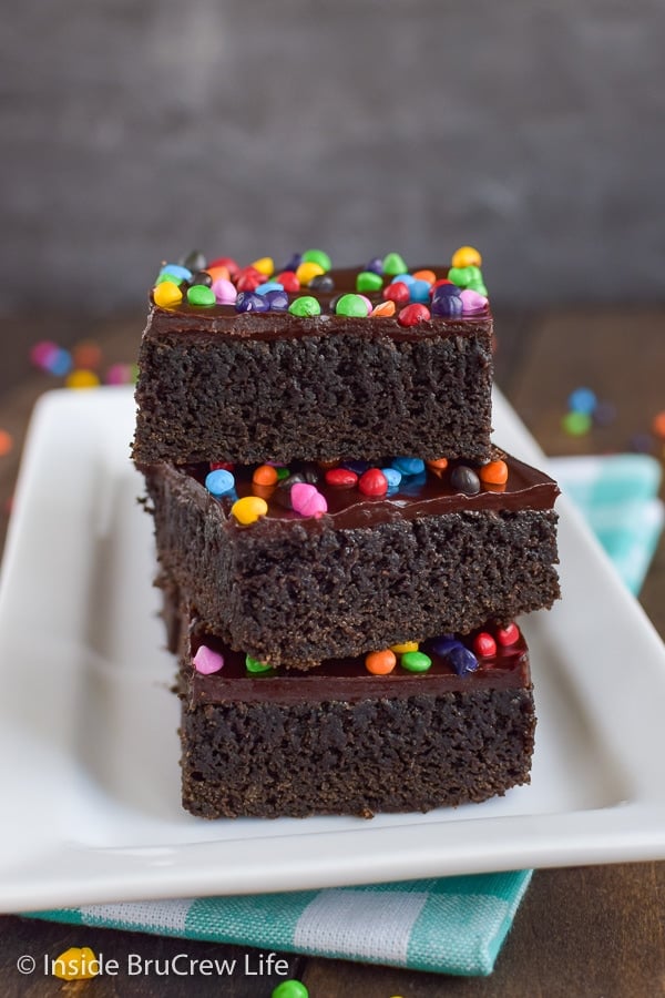 Three chocolate brownies topped with rainbow chips stacked on a white plate.