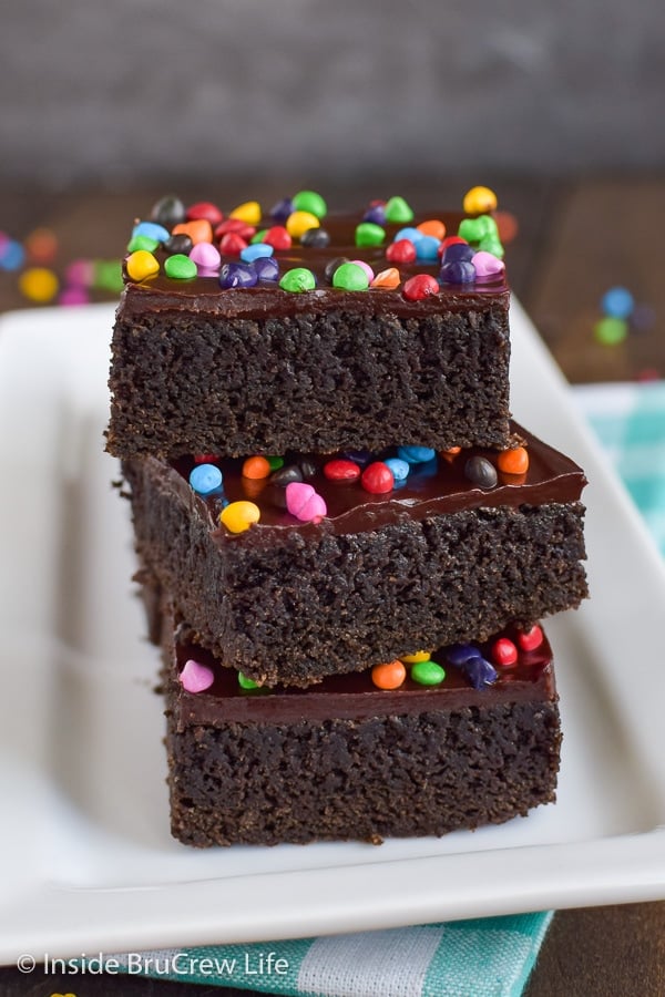 A stack of chocolate brownies topped with frosting and rainbow chips on a white plate.