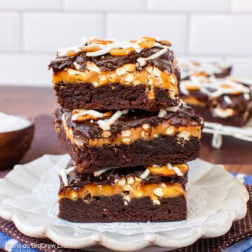 Three chocolate brownies topped with caramel and pretzels stacked on a plate.