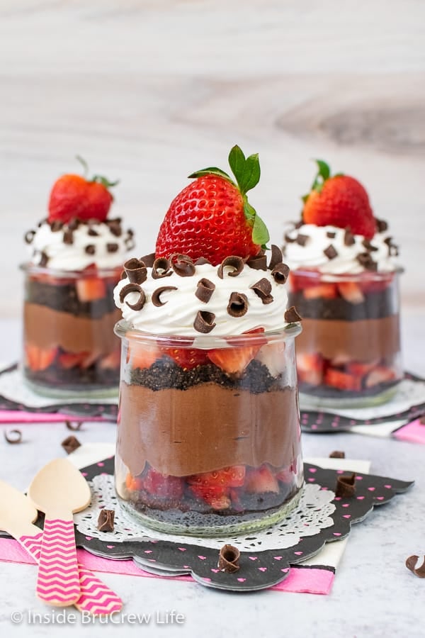 Three jars with layers of no bake chocolate cheesecake, Oreos, and strawberries topped with Cool Whip and a strawberry