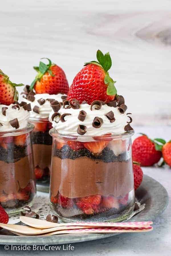 Clear jars of strawberry chocolate cheesecake parfaits topped with Cool Whip and a strawberry on a metal tray