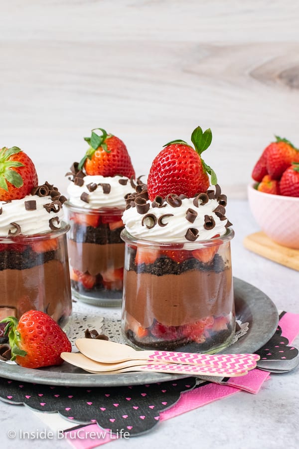 Three clear jars filled with no bake cheesecake, whipped cream, chocolate curls, and fresh strawberries.