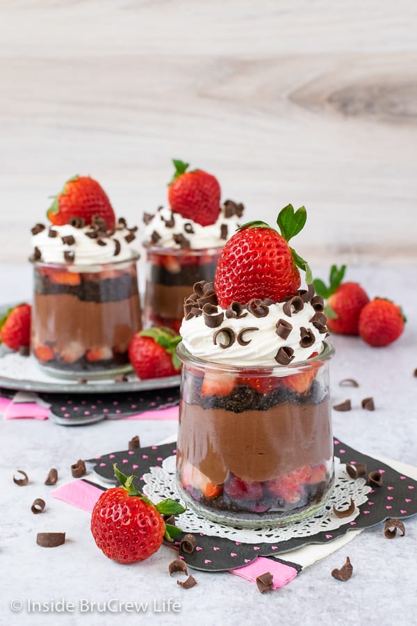 Three clear jars filled with layers of strawberry chocolate cheesecake and topped with whipped topping and fresh strawberries