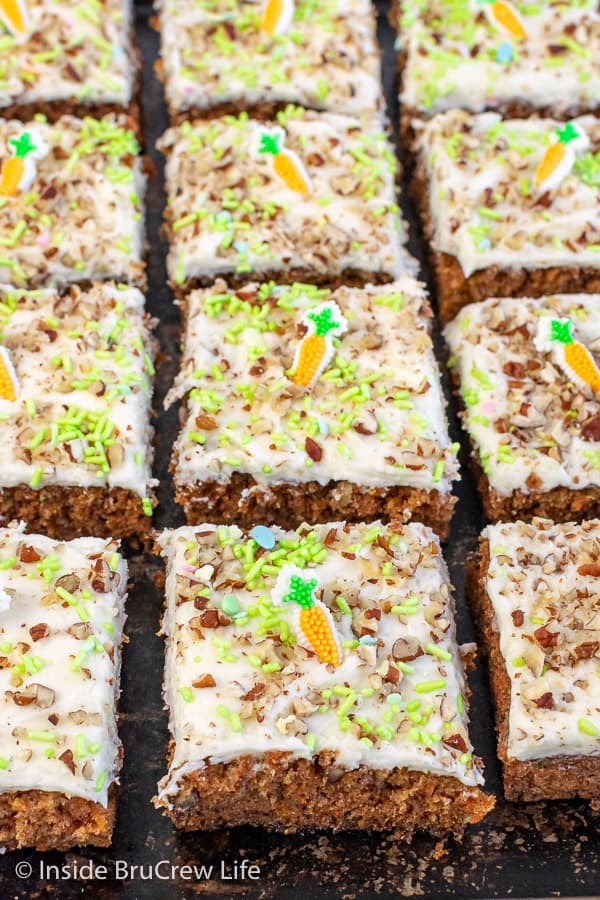 A black sheet pan covered with squares of carrot cake bars topped with frosting, pecans, and green sprinkles.