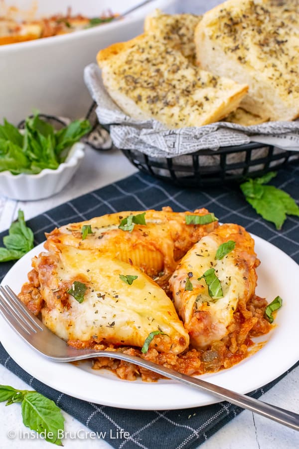 A white plate with three stuffed shells on meat sauce and a basket of garlic bread behind it.