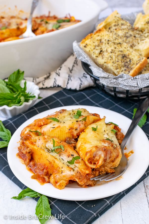 A white plate with three stuffed shells and a fork on it and garlic bread behind it.