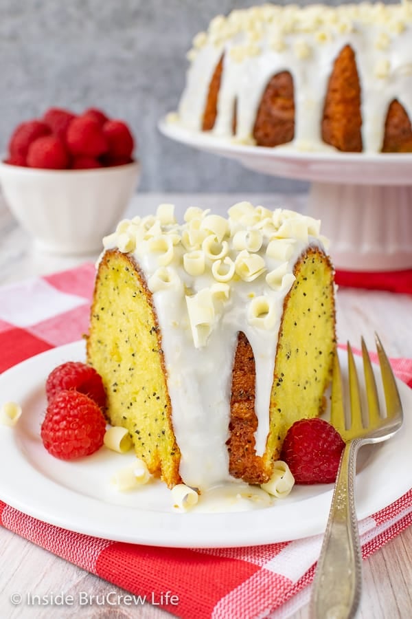 A slice of lemon cake with lemon glaze and fresh raspberries on a white plate with the lemon bundt cake behind it