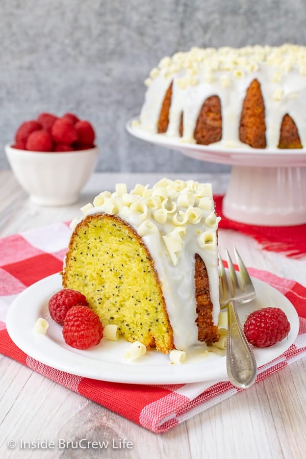 A slice of lemon bundt cake with lemon glaze on a white plate with a bowl of raspberries behind it