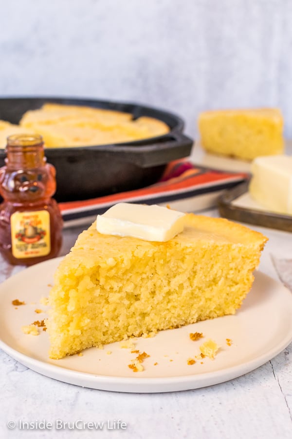 A wedge of easy cornbread with a pat of butter on an off white plate with a skillet of cornbread behind it