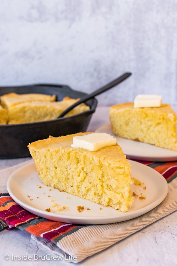 A wedge of easy cornbread on an off white plate with a skillet of cornbread behind it