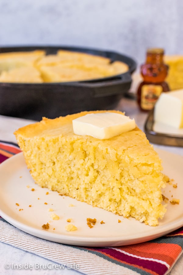 A wedge of skillet cornbread on an off white plate with the skillet behind it