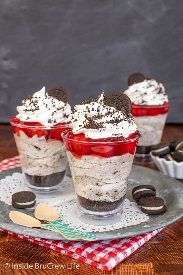 Three clear cups filled with cherry pie filling, cookies and cream cheesecake, and cookie crumbs on a metal tray