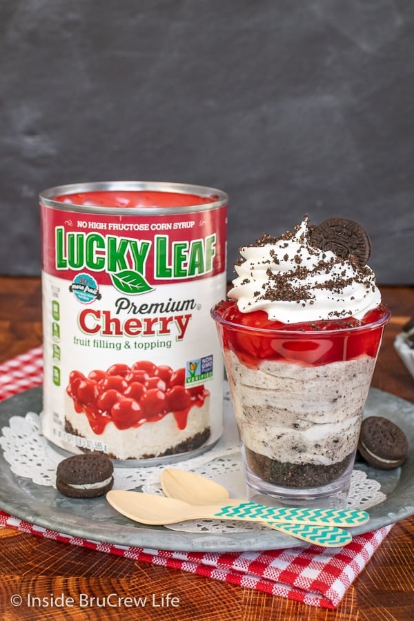 A clear cup with layers of cookies, cookies and cream cheesecake, and cherry pie filling on a metal tray with a can of pie filling beside it