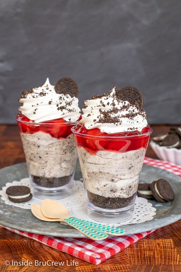 Two clear cups on a metal tray filled with layers of cookie crumbs, cookies and cream cheesecake, and cherry pie filling