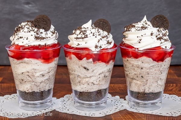 Three clear cups filled with cookie crumbs, cookies and cream cheesecake, and cherry pie filling side by side