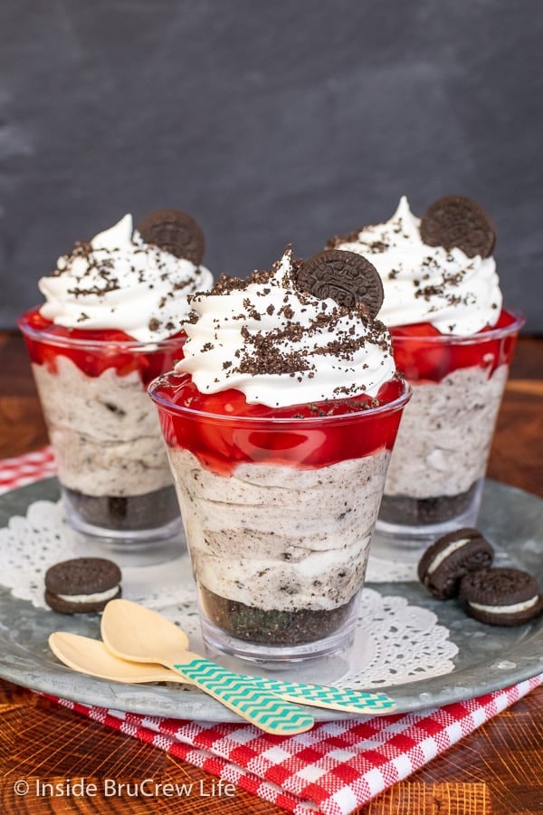 Three parfait cups on a metal tray filled with layers of cookie crumbs, cookies and cream cheesecake, and cherry pie filling