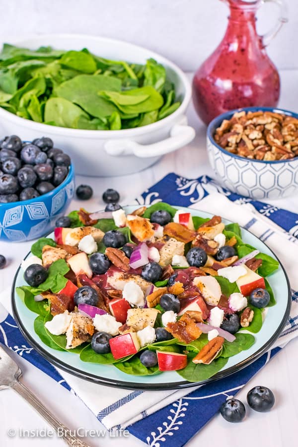 A plate of spinach salad topped with fruit, cheese, nuts, and grilled chicken with bowls of toppings behind it