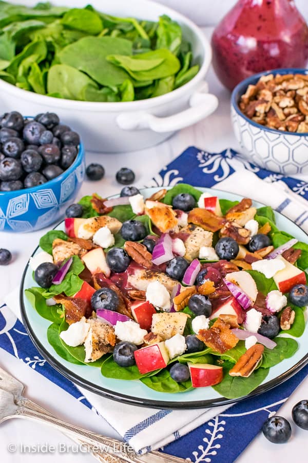 A close up picture of a blue plate with a spinach salad topped with blueberries, cheese, apples, and grilled chicken with bowls of toppings behind it