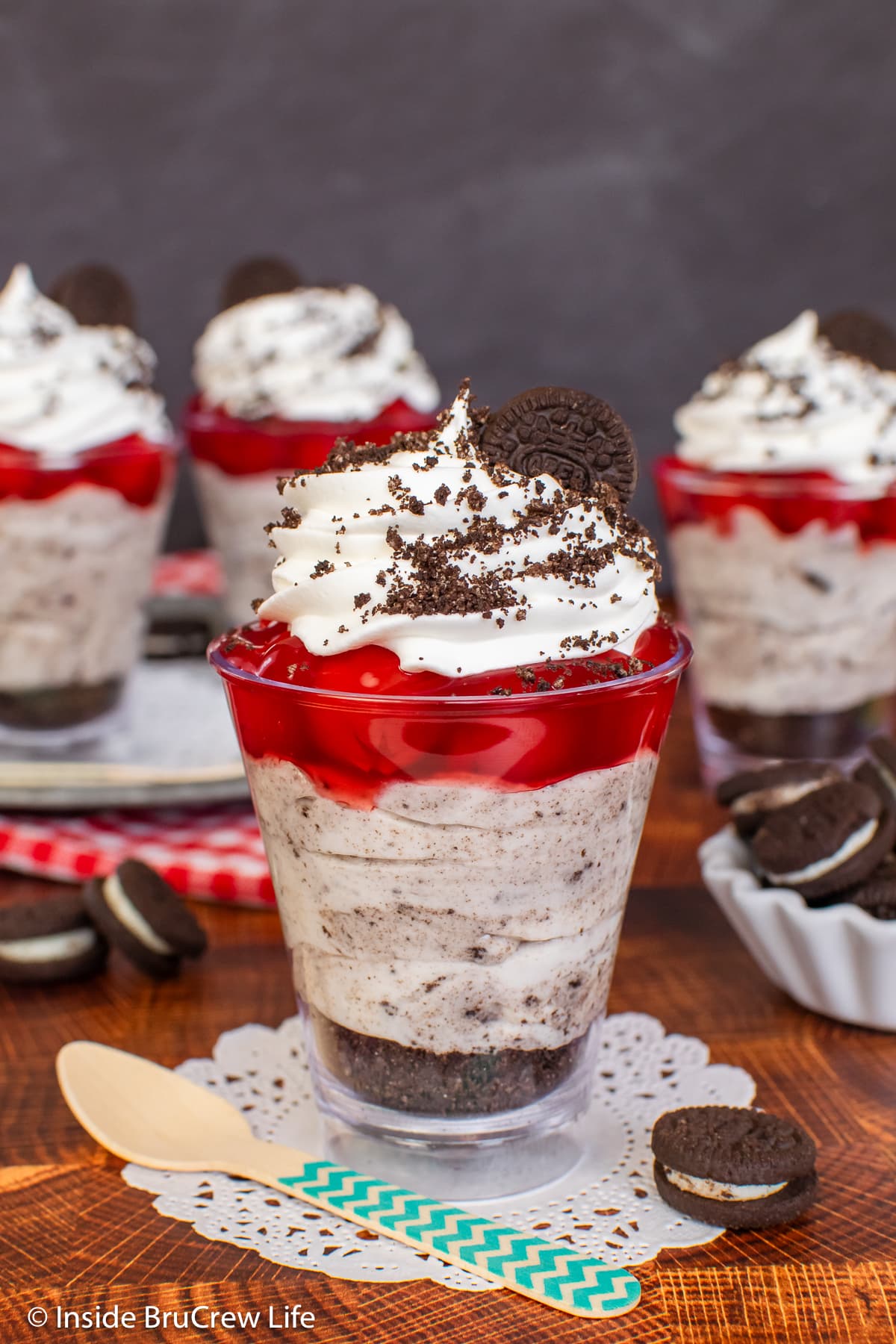 Mini cups of cookies and cream parfaits topped with cherries on a brown board.