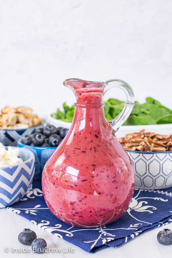 A close up picture of a glass jar filled with Blueberry Balsamic Salad Dressing with bowls of salad ingredients behind it