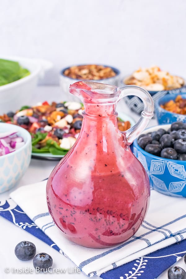 A glass jar filled with Creamy Blueberry Balsamic Salad Dressing with bowls of salad ingredients behind it