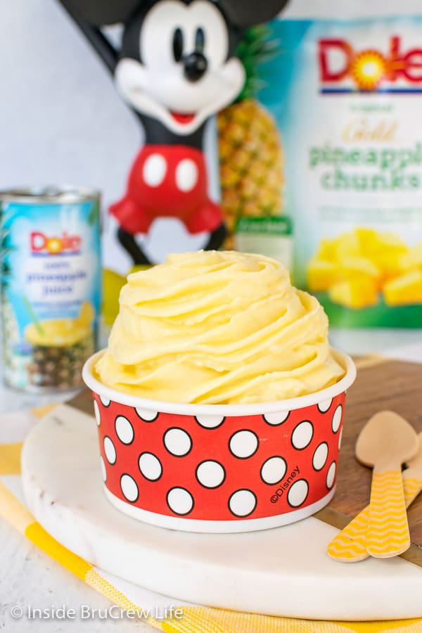 A red and white polkadot cup filled with a swirl of pineapple Dole Whip with pineapple juice and chunks behind it
