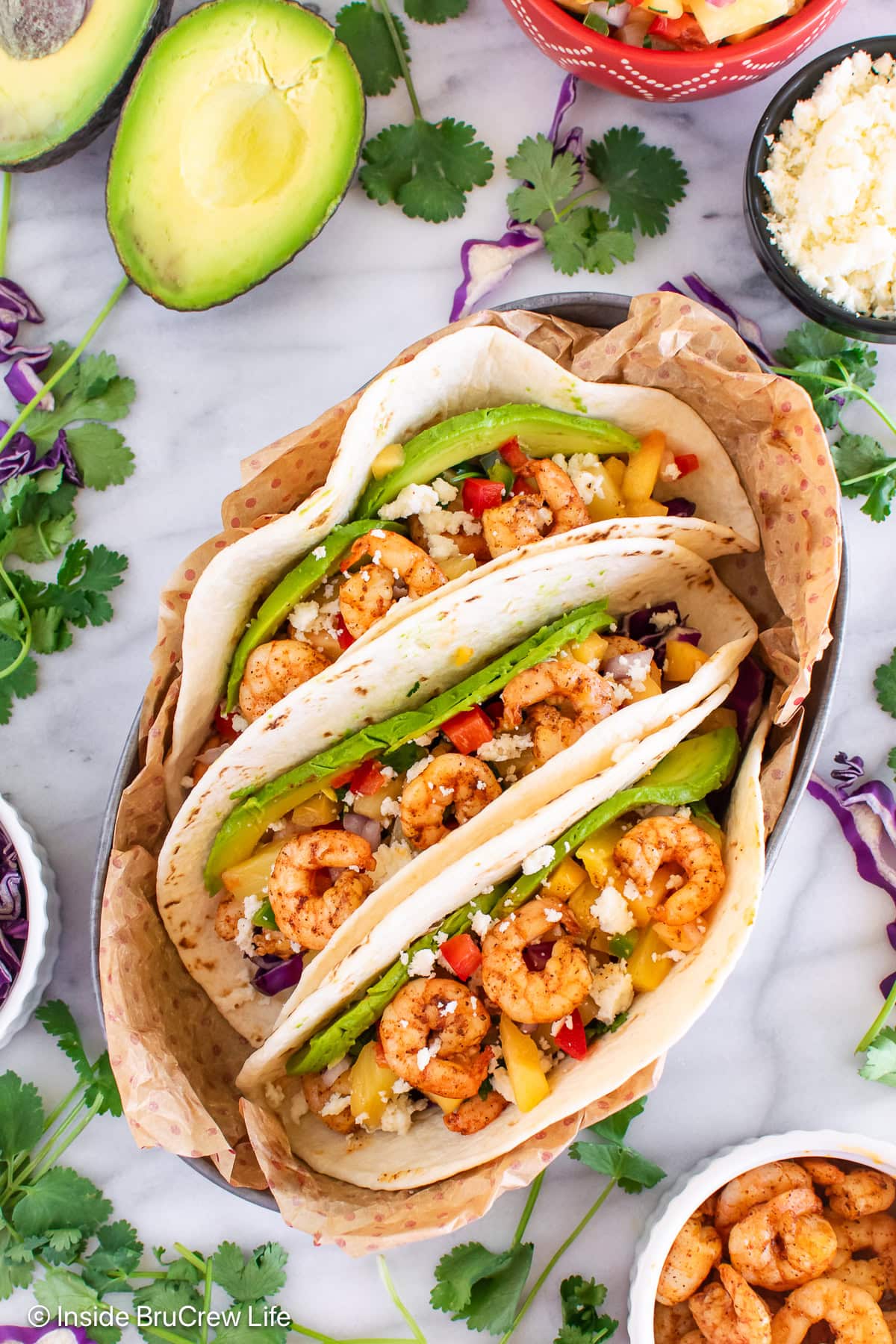 Three shrimp tacos with toppings in a metal serving dish.