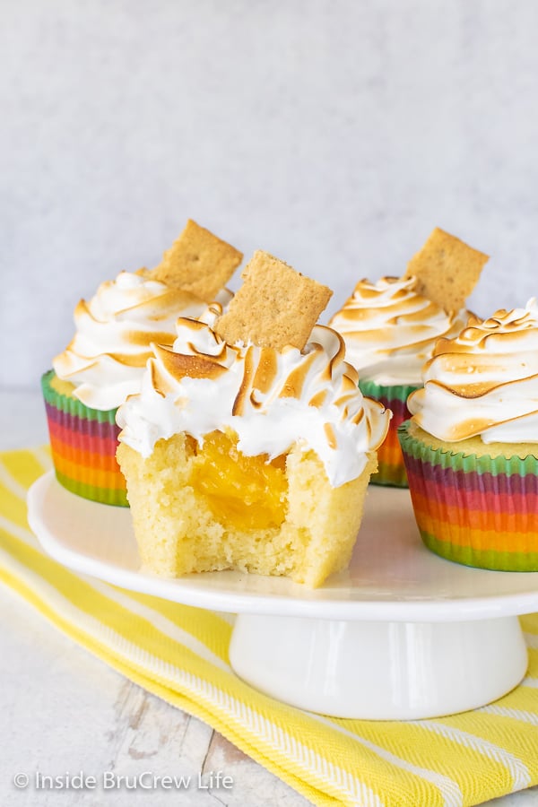 Four Lemon Meringue Cupcakes on a white cake plate with a yellow towel under it. One cupcake has a bite out of it showing the hidden lemon center.