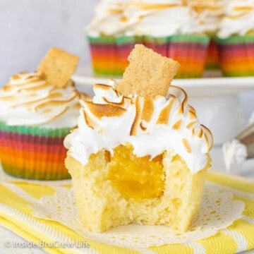 A close up picture of a lemon meringue cupcake on a white doily and yellow towel with a bite take out to show the hidden lemon center